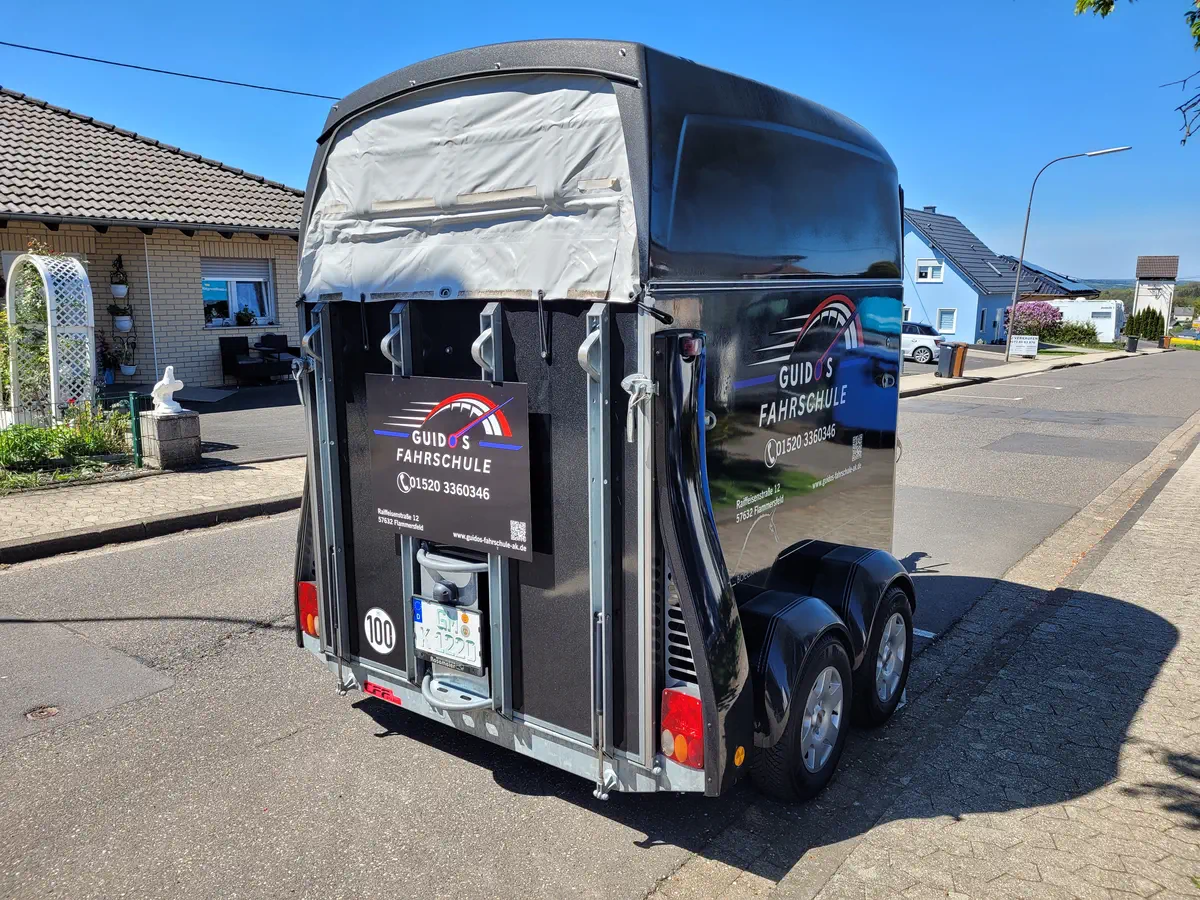 Heckansicht eines Anhängers mit auffälliger Fahrzeugbeschriftung für "Guido's Fahrschule", inklusive Kontaktdaten und Logo.